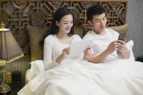 Coppia cinese che utilizza tablet digitale e smartphone a letto — Foto stock