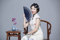 Mujer china en vestido tradicional sentada en la mesa de té y sosteniendo ventilador de mano - foto de stock