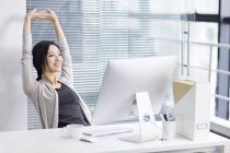 Mujer china estirando los brazos en la oficina y mirando a través de la ventana - foto de stock