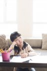 Menina chinesa se apoiando no braço e olhando para longe ao fazer lição de casa — Fotografia de Stock