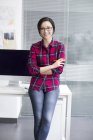 Mujer china apoyada en la mesa con los brazos cruzados en la oficina - foto de stock
