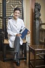 Médica chinesa sentada na cadeira e segurando diário — Fotografia de Stock