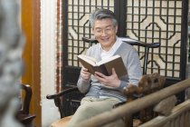Hombre chino mayor sentado en silla y libro de lectura - foto de stock