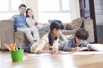 Chinese siblings studying together at floor with parents on sofa watching — Stock Photo