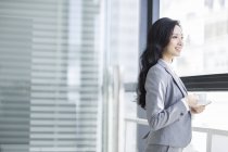 Chinese businesswoman holding coffee and looking through window at office — Stock Photo