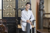 Mujer china doctor sentado en silla y mirando en cámara - foto de stock