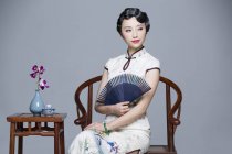 Mujer china en vestido tradicional sentada en la mesa de té y sosteniendo ventilador de mano - foto de stock