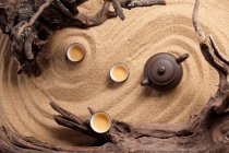 Vista de panela de chá e copos na superfície de areia com madeira à deriva — Fotografia de Stock