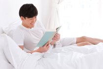 Young Chinese man reading a book in bed — Stock Photo