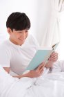 Jovem chinês lendo um livro na cama — Fotografia de Stock