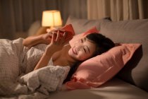 Young chinese woman using smartphone in bed — Stock Photo