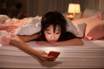 Mujer china joven usando teléfono inteligente en la cama - foto de stock