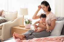 Müde Mutter hält Baby auf Couch mit der Hand am Kopf — Stockfoto