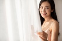 Glückliche junge Chinesin lächelt in die Kamera, während sie am Fenster mit Vorhängen steht — Stockfoto