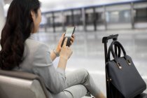 Junge Chinesin benutzt Smartphone während sie im Flughafen sitzt — Stockfoto