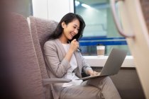 Mujer sonriente sentada en tren de alta velocidad con taza de café y el uso de ordenador portátil - foto de stock