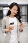 Young chinese woman holding coffee and using smartphone in airport — Stock Photo