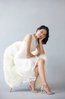 Красивая китаянка позирует на пушистом кресле в белом платье и на высоких каблуках — стоковое фото