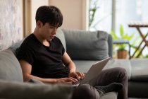 Jeune homme chinois en utilisant un ordinateur portable assis sur le canapé — Photo de stock