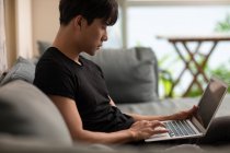 Молодий китайський чоловік використовує ноутбук, сидячи на дивані — стокове фото