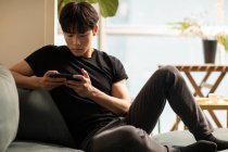 Junger Chinese benutzt Smartphone, während er auf der Couch sitzt — Stockfoto