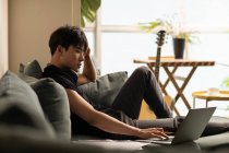 Jovem chinês homem usando laptop sentado no sofá — Fotografia de Stock