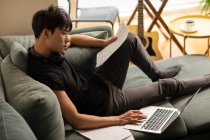 Молодий китайський чоловік використовує ноутбук з паперами на дивані — стокове фото