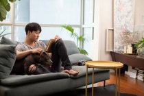 Jovem chinês homem com cão sentado no sofá e livro de leitura — Fotografia de Stock