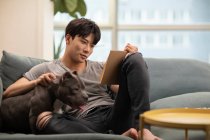 Jovem chinês homem com cão sentado no sofá e livro de leitura — Fotografia de Stock