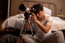 Young chinese man looking through telescope sitting beside bed — Stock Photo