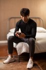 Молодий китайський чоловік з навушниками, дивлячись на екран смартфона, сидячи на ліжку — стокове фото