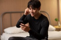 Усміхнений китайський чоловік дивиться на екран смартфона і використовує навушники — стокове фото