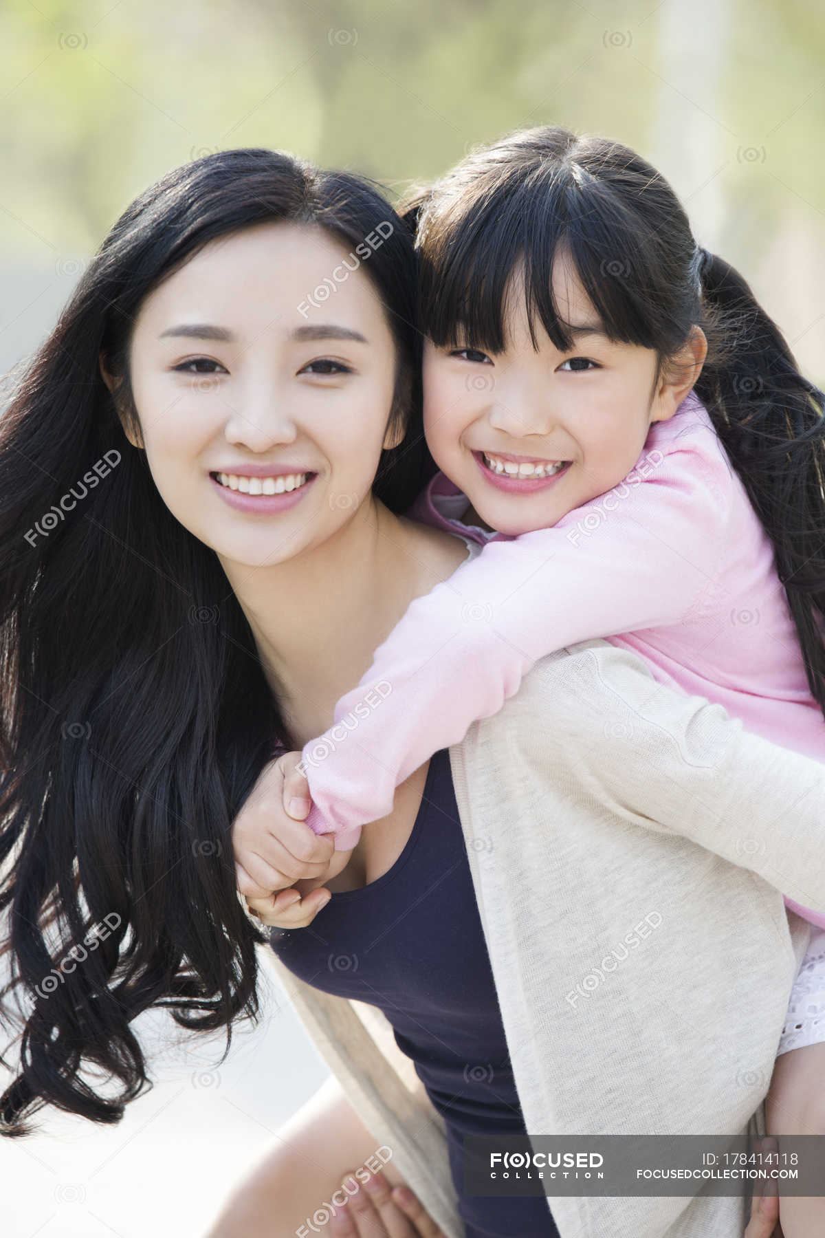 Asian mother riding daughter piggyback — outside, cheerful 