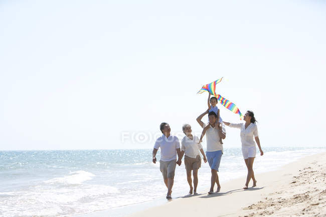 Menina chinesa com pipa nos ombros do pai andando com a família na praia — Fotografia de Stock
