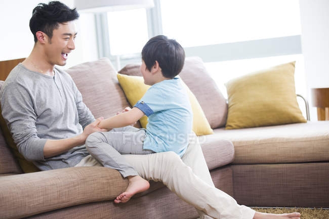 Padre e hijo chinos jugando y tomados de la mano en el sofá - foto de stock