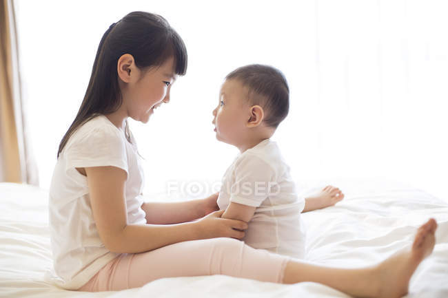 Chinese girl and baby boy sitting face to face on bed — Stock Photo