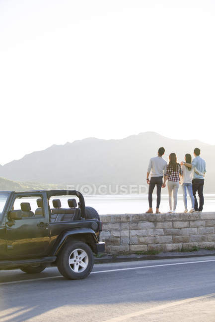Rear view of friends standing on lakeside in suburbs with car — Stock Photo