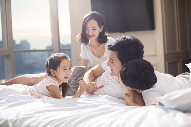 Padres chinos con niños relajándose y divirtiéndose en la cama - foto de stock