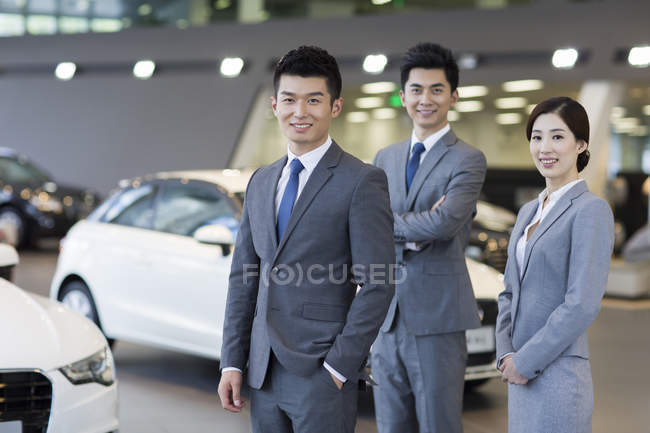 Confident salespeople standing with new cars in showroom — Stock Photo