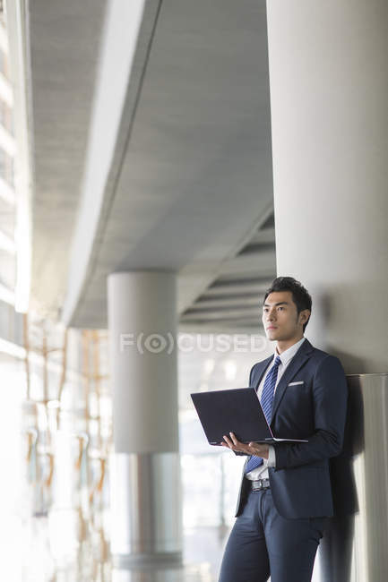 Uomo d'affari cinese in piedi con computer portatile al chiuso — Foto stock