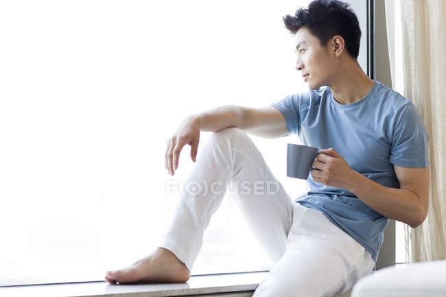 Китаец держит кофе и смотрит в окно дома — стоковое фото