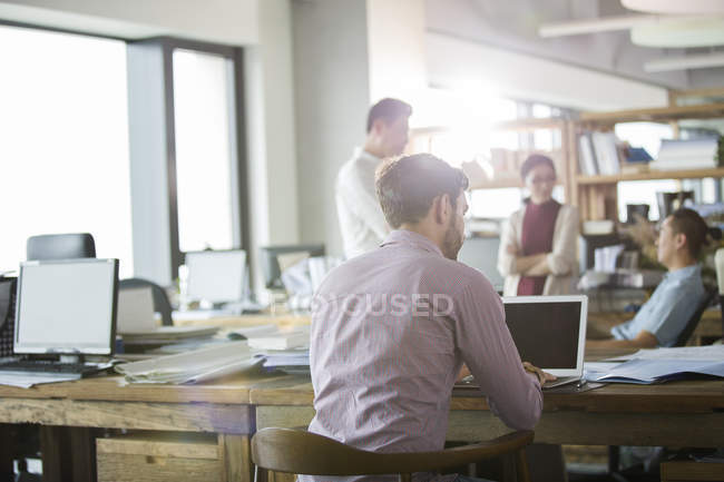 Rear view of office worker using laptop with colleagues in background — Stock Photo