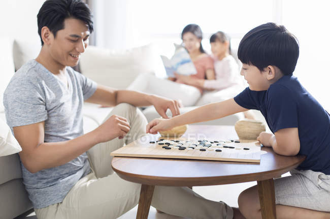 Menino chinês jogando jogo de Ir com o pai, enquanto a mãe ea filha lendo em segundo plano — Fotografia de Stock