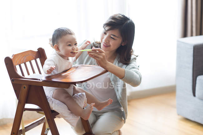 Chinese mother feeding baby boy in high chair — Stock Photo