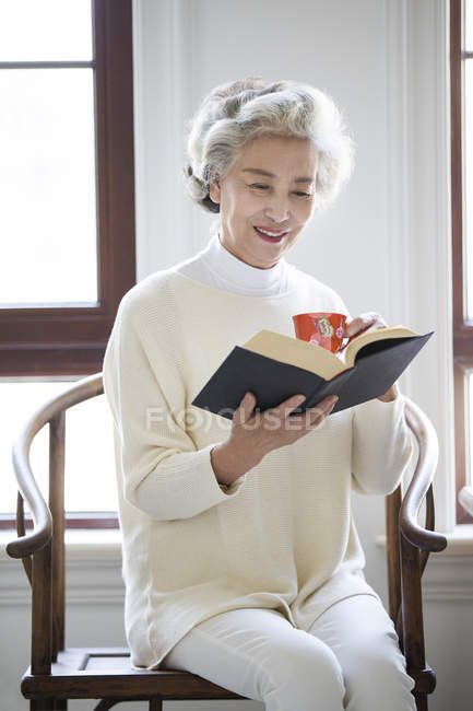 Senior Chinese woman reading book with cup of tea — Stock Photo