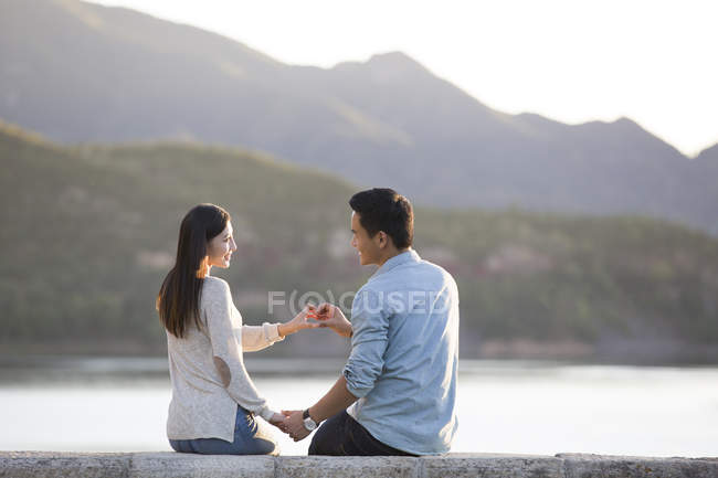 Chinese couple sitting on lakeside in suburbs and making love sign with hands — Stock Photo