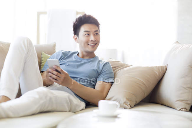 Hombre chino sosteniendo teléfono inteligente y mirando hacia otro lado en el sofá - foto de stock