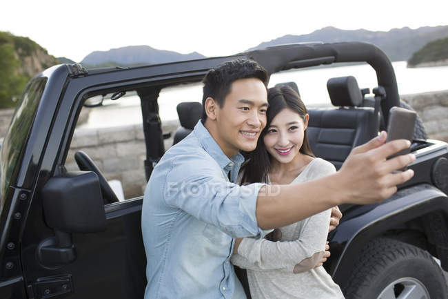 Pareja china tomando selfie con smartphone delante del coche - foto de stock
