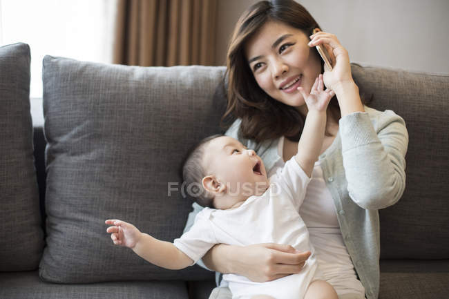 Chinês mãe falando no telefone enquanto segurando menino em casa — Fotografia de Stock