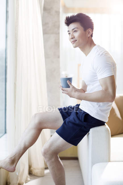Chinese man holding coffee and looking through window at home — Stock Photo
